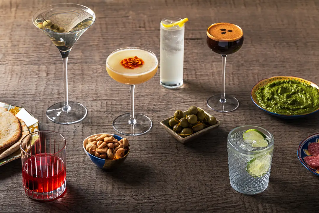 Cocktails served with small food pairings on a dark table, illustrating how drinks and dishes complement each other.