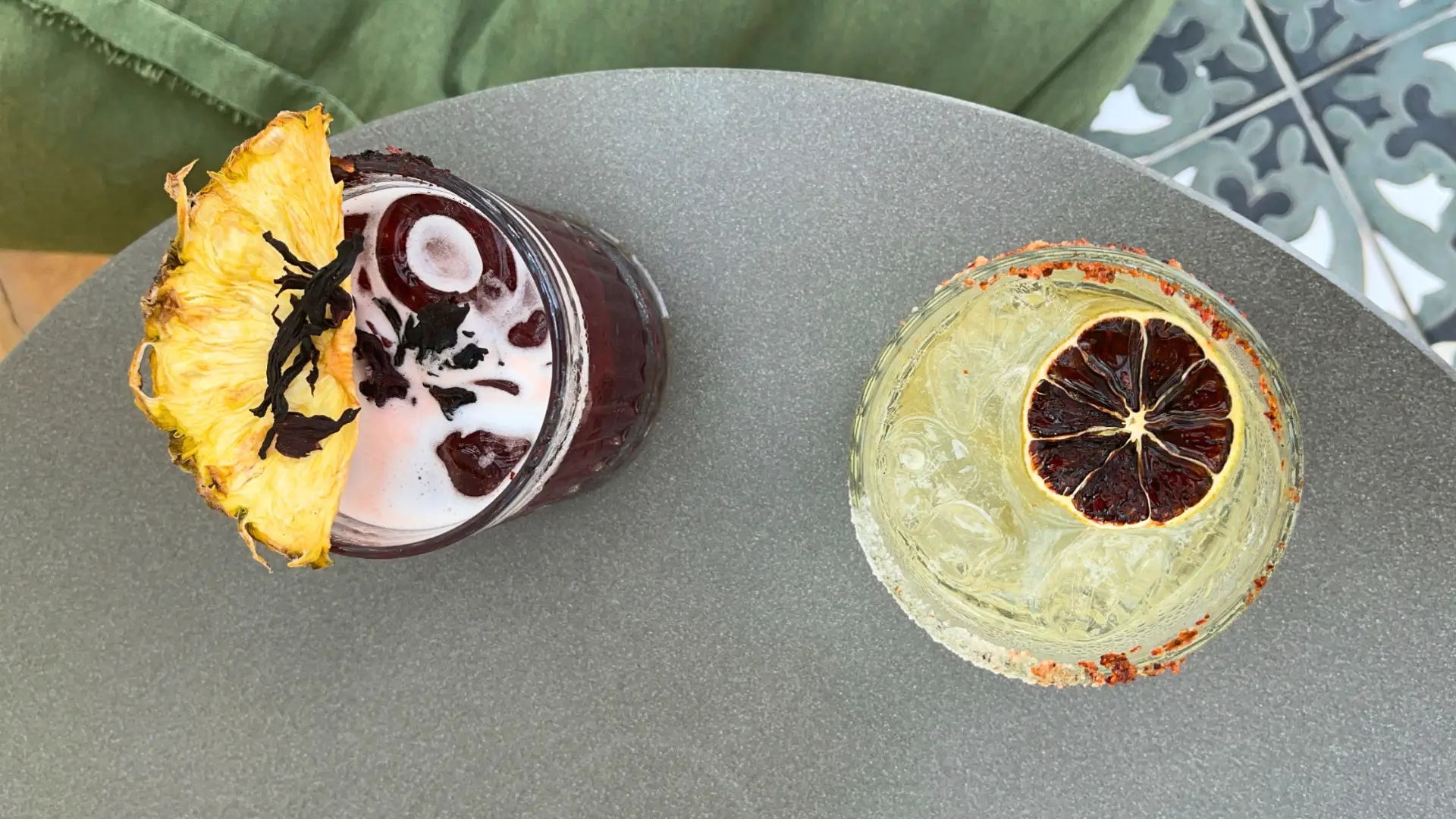 Two cocktails with sustainable garnishes: one topped with a dried pineapple slice and hibiscus, the other with a dehydrated citrus wheel and spiced rim.