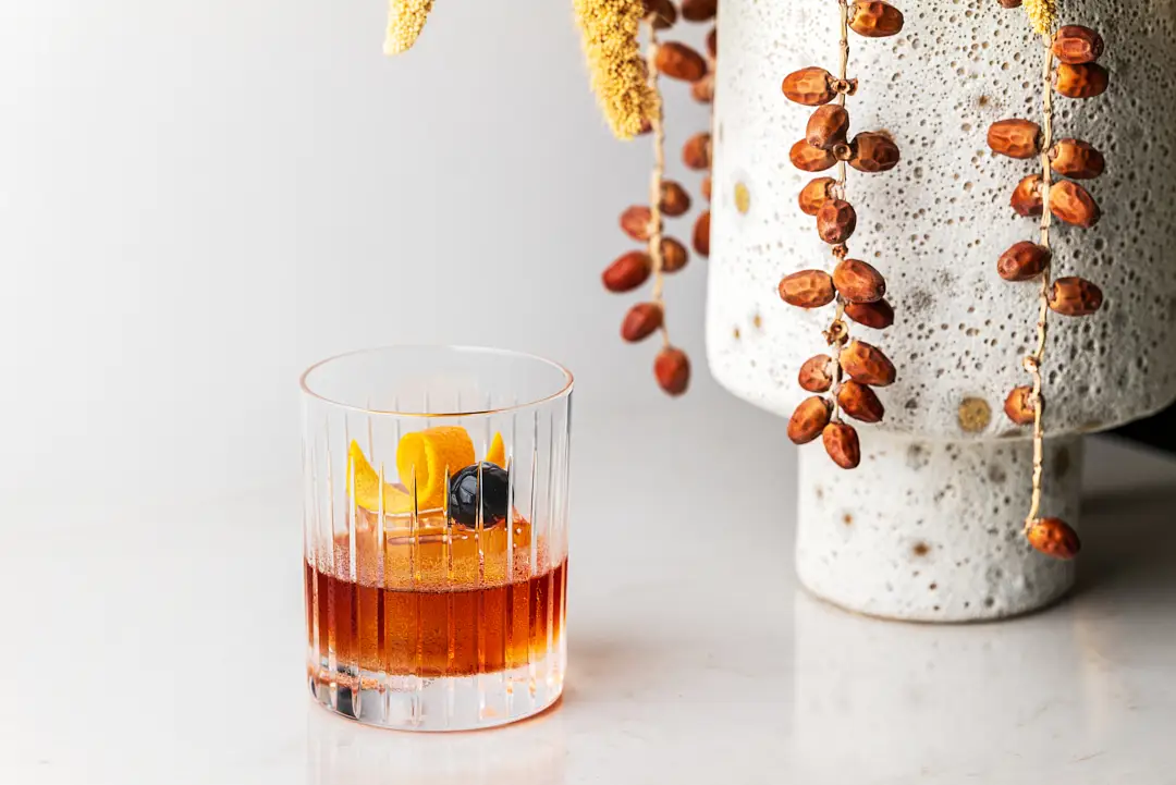 Old Fashioned cocktail in a patterned rocks glass with orange twist and cherry garnish, styled next to a ceramic vase with dried pods.